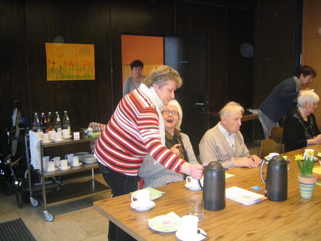 Betreuungsgruppe: Ältere Menschen trinken Kaffee, serviert von einer Frau in gestreiftem Oberteil.