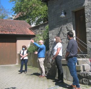Four people wearing masks stand outside a stone building, one gesturing with his arm.