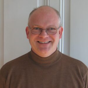 Portrait of a man with glasses in a brown turtleneck sweater smiling.